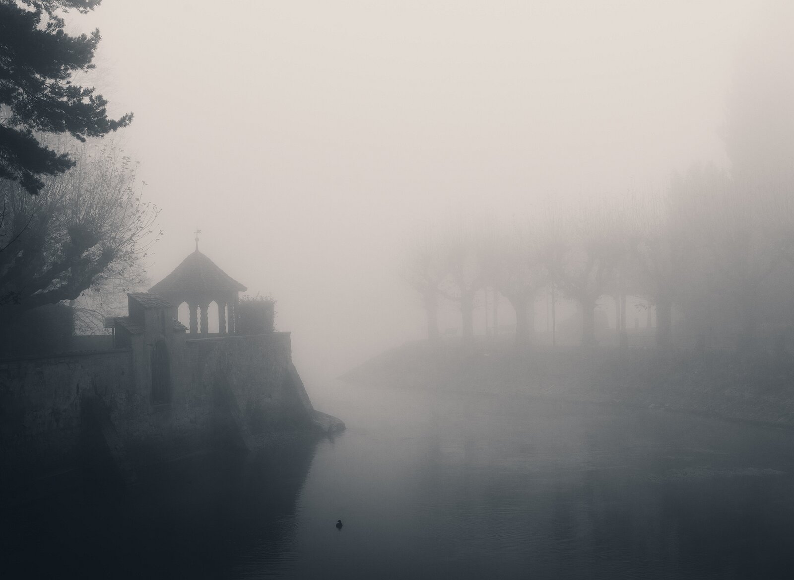 Fog Chapel Small stone riverside structure and nearby trees dissolve into dense mist over still water.