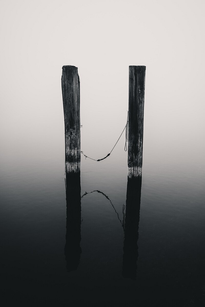 Tethered Quiet Two weathered wooden posts stand in calm water with a rope loosely connecting them, reflected in the still surface.
