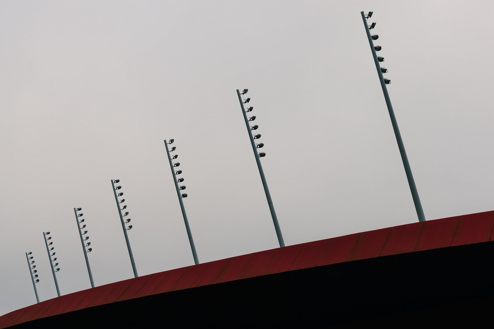 Linear Illumination Rows of slender stadium light poles angle upward above a red roof beneath a pale sky.