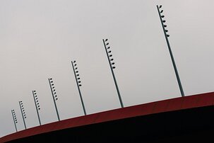 Linear Illumination Rows of slender stadium light poles angle upward above a red roof beneath a pale sky.