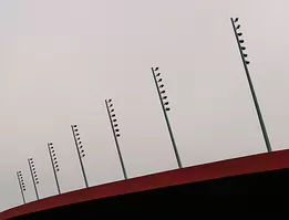 Measured Ascent Minimalist architectural photograph featuring a line of stadium floodlights ascending above a red roof against a soft grey sky - a study in balance, rhythm, and...
