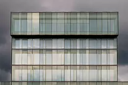 Stark Modernism Tall, symmetrical glass building reflecting soft light against a backdrop of dark clouds.