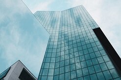 Reflective Heights Blue-tinted glass tower seen from ground level, its façade reflecting clouds and sky.