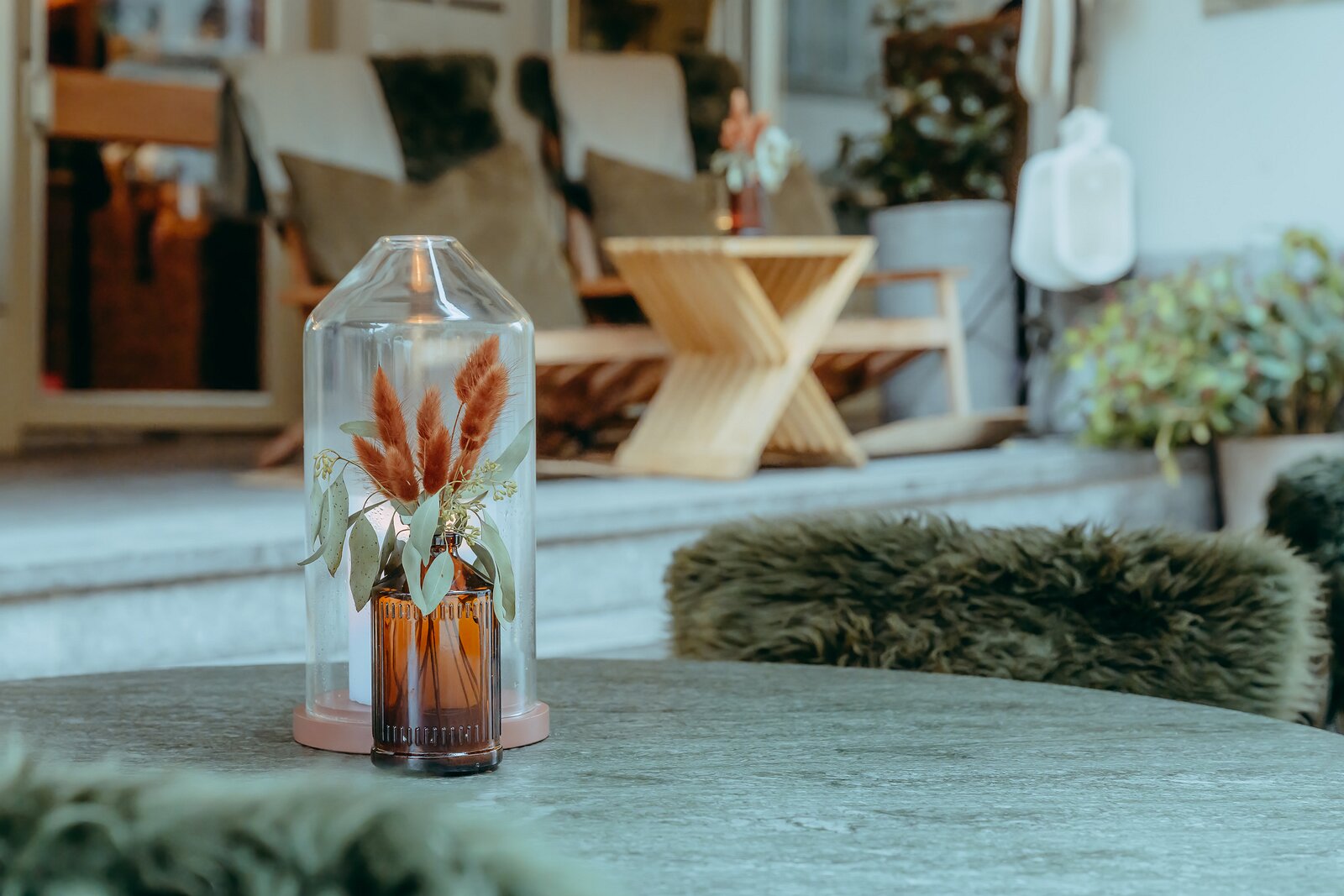 Cozy Still A dried floral arrangement in an amber bottle under a glass dome sits on a round table in a cozy outdoor seating area.