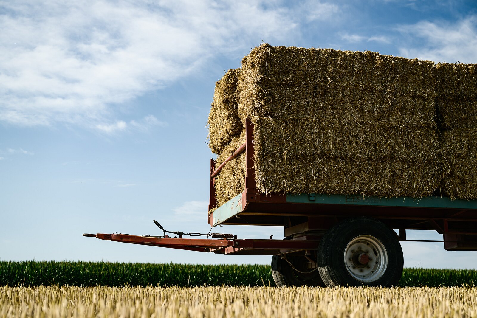 Harvest Still Farm trailer stacked high with square hay bales sits in a harvested field under a bright blue sky.