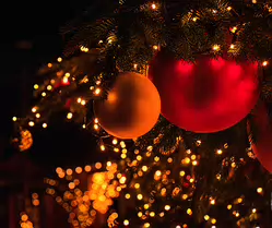 Ember of Winter Warm close-up of red and gold Christmas ornaments hanging from pine branches, illuminated by soft golden fairy lights with rich bokeh in the background.