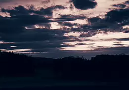 Fading Quietude A moody twilight landscape captured near Frauenfeld, Switzerland, featuring dramatic layered clouds and a subtle evening glow above a silhouetted treeline. The...