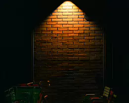 Under the Lamp A lone overhead lamp casts a warm cone of light onto a brick wall, revealing a bicycle and a row of empty chairs fading into shadow. A quiet urban moment where...