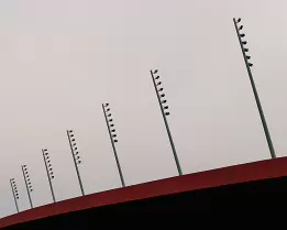 Measured Ascent Minimalist architectural photograph featuring a line of stadium floodlights ascending above a red roof against a soft grey sky - a study in balance, rhythm, and...