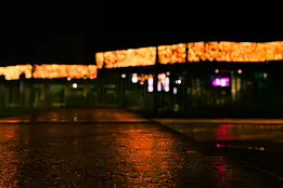 Glowline Low-angle night photograph showing warm orange holiday lights blurred into a horizontal ribbon above a wet pavement, with soft neon reflections in pink and...