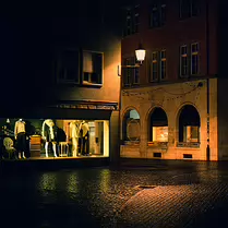 The Still Hour A quiet town square after dark, illuminated by a shop window and a solitary street lamp. Rain-soaked cobblestones reflect the warm glow, turning an empty scene...