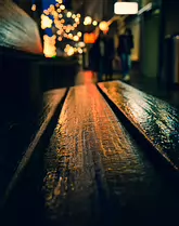 Where the Night Rests Rain-darkened wood catches the glow of distant streetlights, turning a simple bench into a quiet stage for the city’s passing life. Soft bokeh and silhouettes...