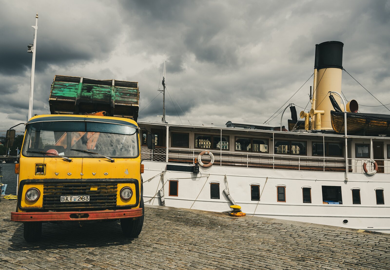 Quayside Cargo An old yellow Volvo truck loaded with crates stands beside a heritage steamboat on a cobbled quay under brooding skies.