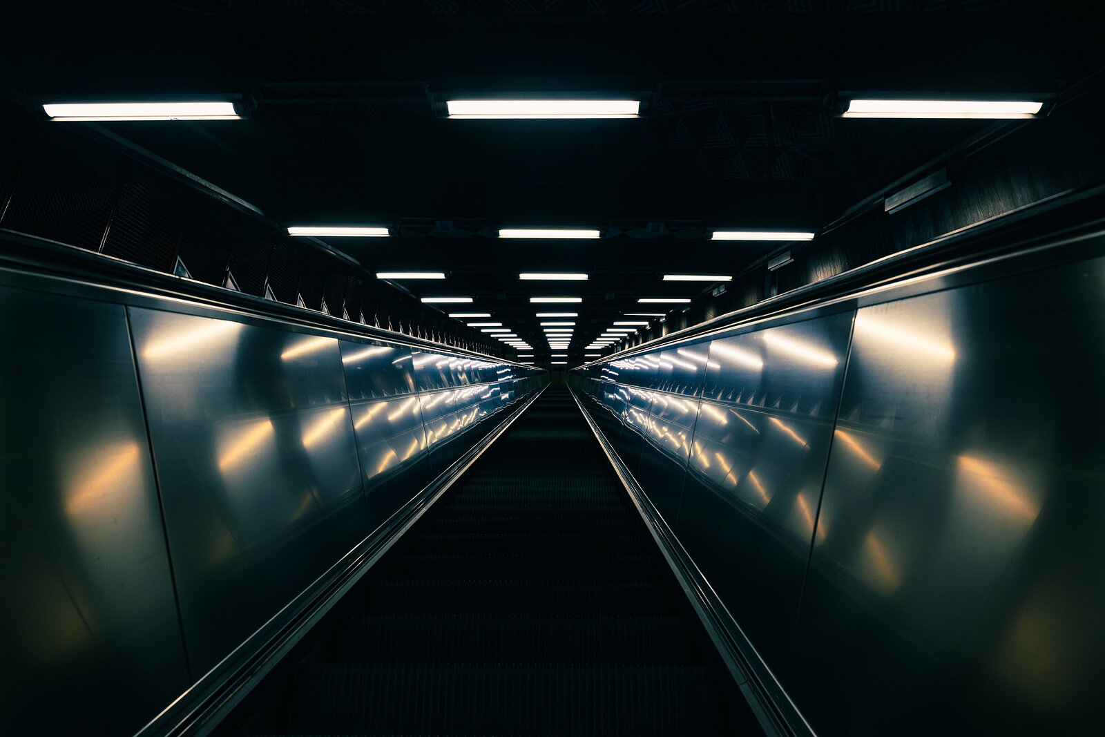Tunnelbana Rise A long, lit escalator tunnel in Stockholm’s metro converges to a vanishing point deep underground.