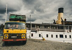 Quayside Cargo An old yellow Volvo truck loaded with crates stands beside a heritage steamboat on a cobbled quay under brooding skies.