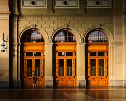 Warm Portals A lone man walks past ornate wooden double doors framed by stone arches, illuminated by warm dawn light.