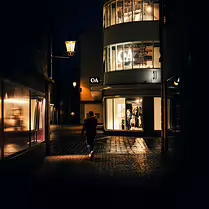 Crossing the Quiet Glow A moody night-street photograph capturing a solitary figure walking past softly illuminated storefronts and cobblestone reflections. The warm streetlights and...