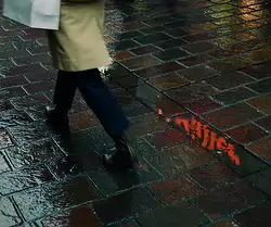 Passing Glow A passerby crosses wet cobblestones as a flicker of red neon ripples through a shallow puddle - an ordinary moment turned quietly cinematic.