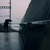 Cold Passage A lone figure drifts through the rain, framed by cold concrete and the gleam of damp metal - an understated moment of quiet transit.