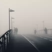 Fog Crossing Pedestrians and a cyclist cross a fog-shrouded bridge lined with evenly spaced lamps.