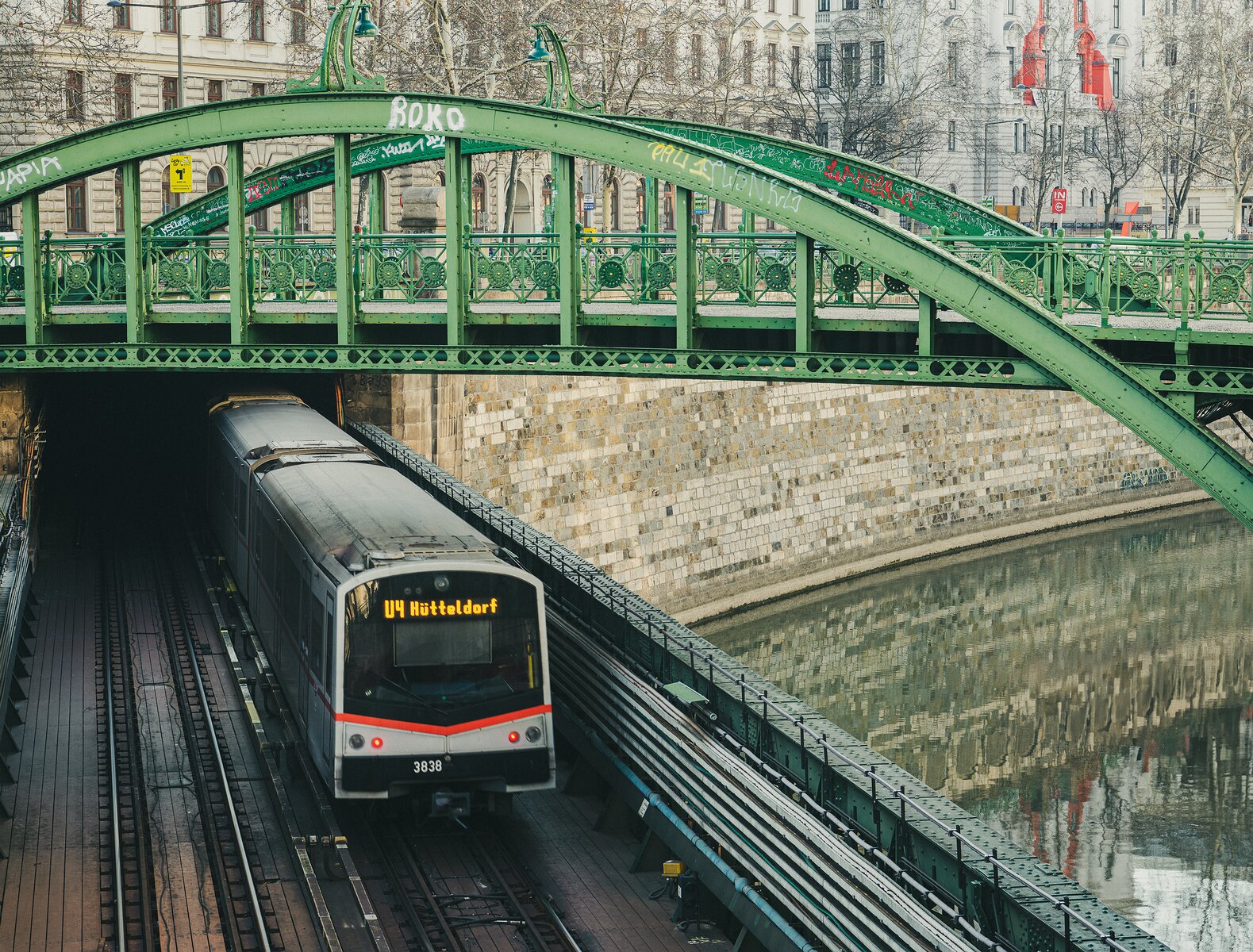 Historic Span Vienna U4 metro train emerging from a tunnel beneath an ornate green iron bridge along the Wien River.