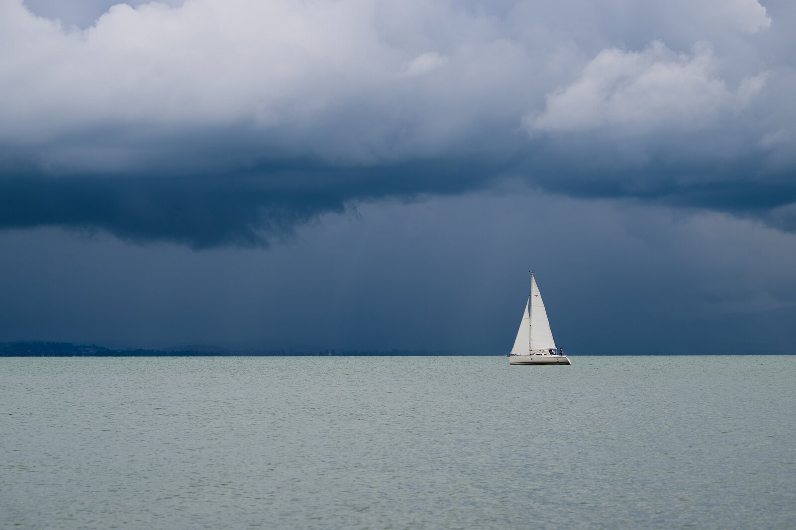 Bold Voyage A lone sailboat floats on still water beneath a looming dark storm front.