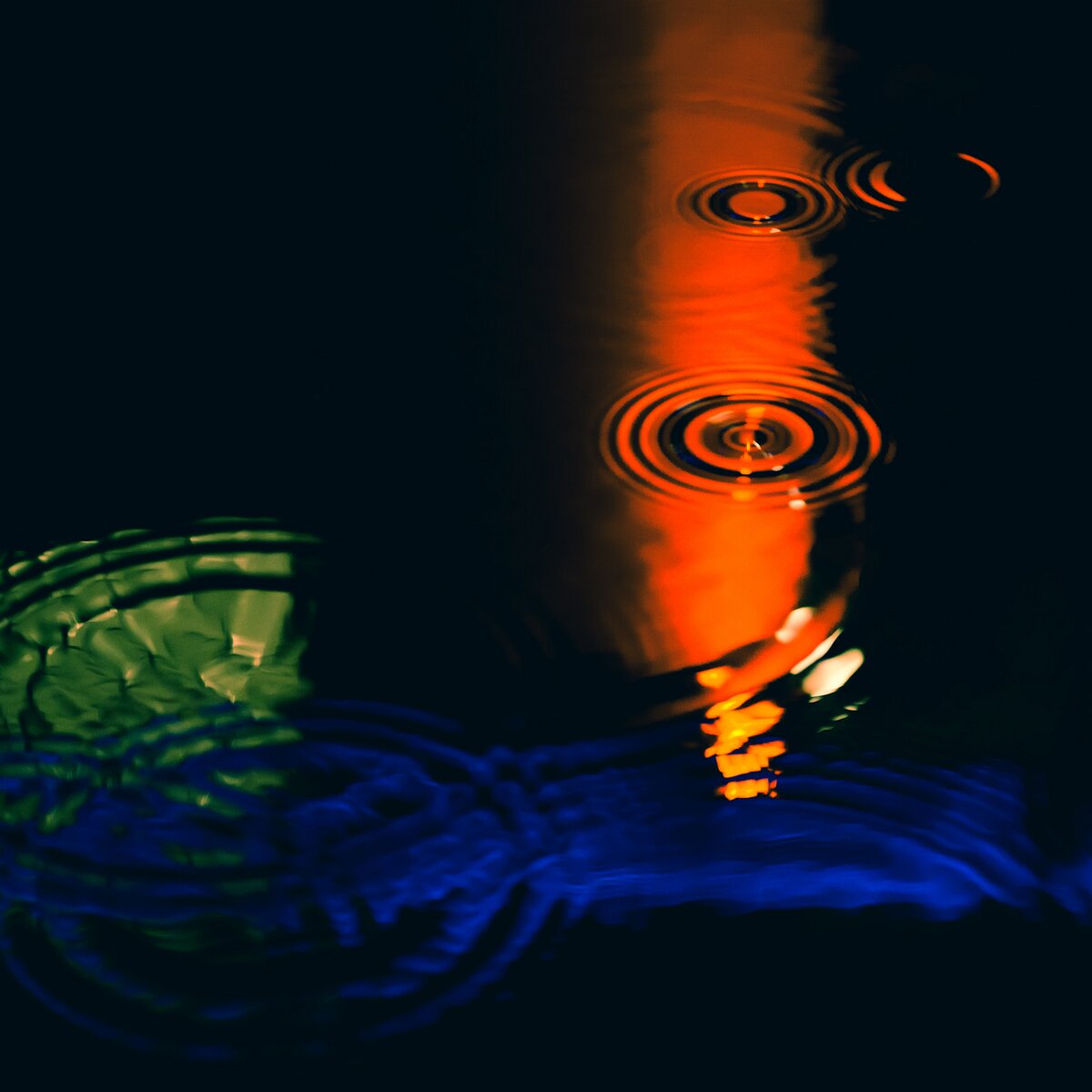 Color Echoes Rippling water reflects distorted streaks of green, blue, and orange neon lights.